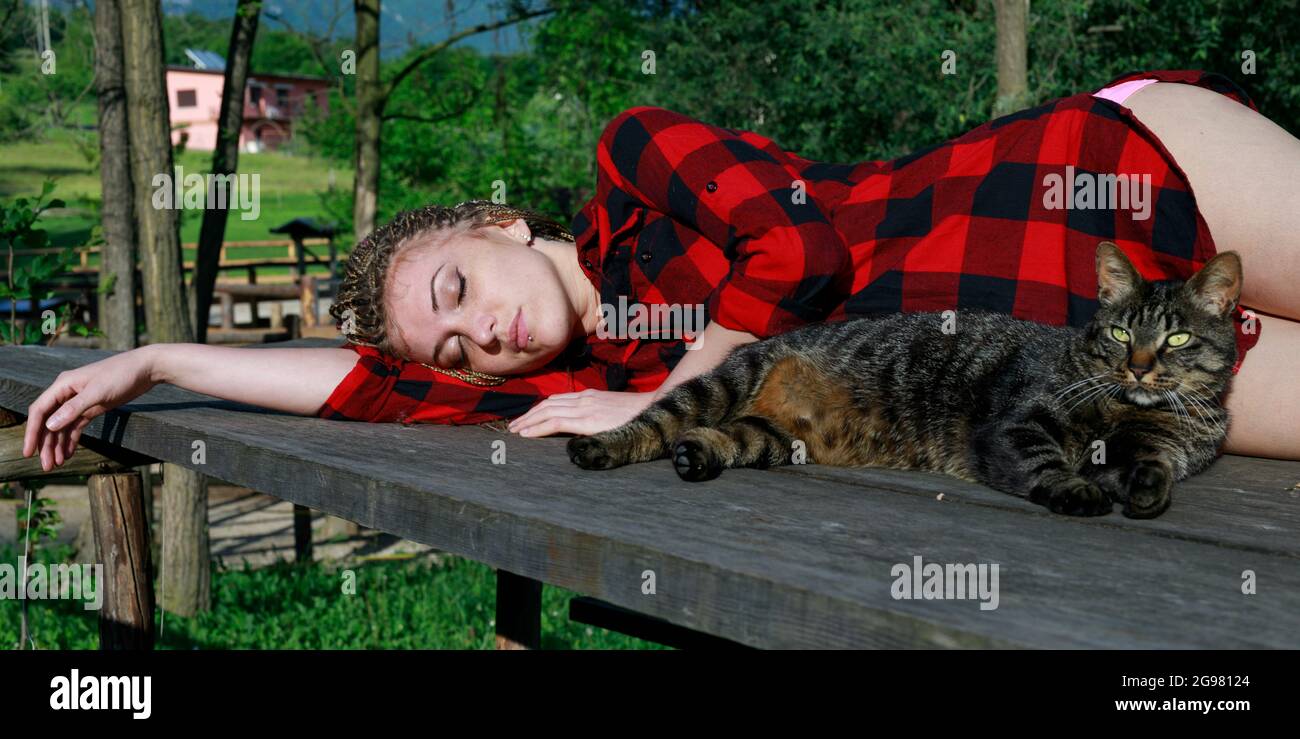 gorgeous woman with braided hairstyle, languidly lying with her eyes closed on the table, just like the cat who is with her in the same attitude Stock Photo