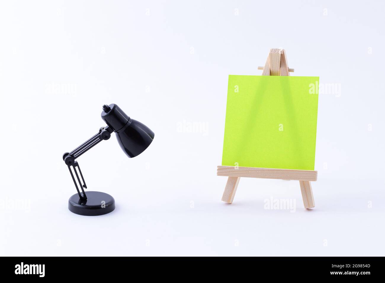 Wooden Easel Miniature with Blank Colored Square Canvas or Memo Paper - Mockup. Mini Wooden Stand with Clean Artboard and Small Black Table Lamp on White Background, Copy Space Stock Photo