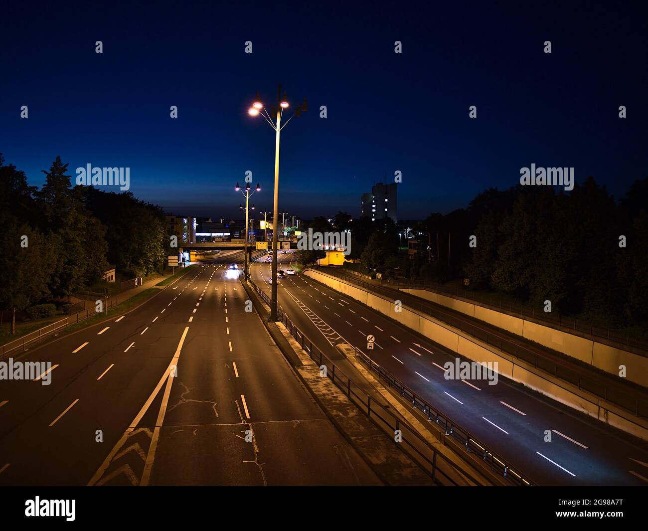 View of main road B9 leading through the center of Koblenz illuminated by lanterns with yellow light and cars passing by at night with trees. Stock Photo
