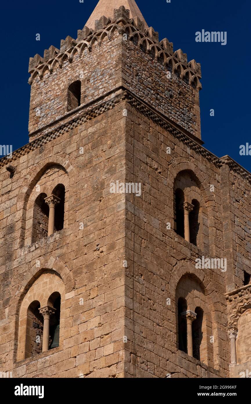 One of the Norman bell-towers soaring above the Duomo di Cefalù, founded in 1131 at Cefalu, Sicily, Italy.  The towers' walls are built of massive cut stone blocks pierced by Romanesque mullioned windows and underline the cathedral's resemblance to a fortress.   The northern belfry, seen here, is surmounted by a smaller rubble tower topped by interlaced arches and swallow-tail merlons, as found on many medieval Italian castles.  Above the rubble tower is an octagonal pyramid spire, added in the 1400s. Stock Photo