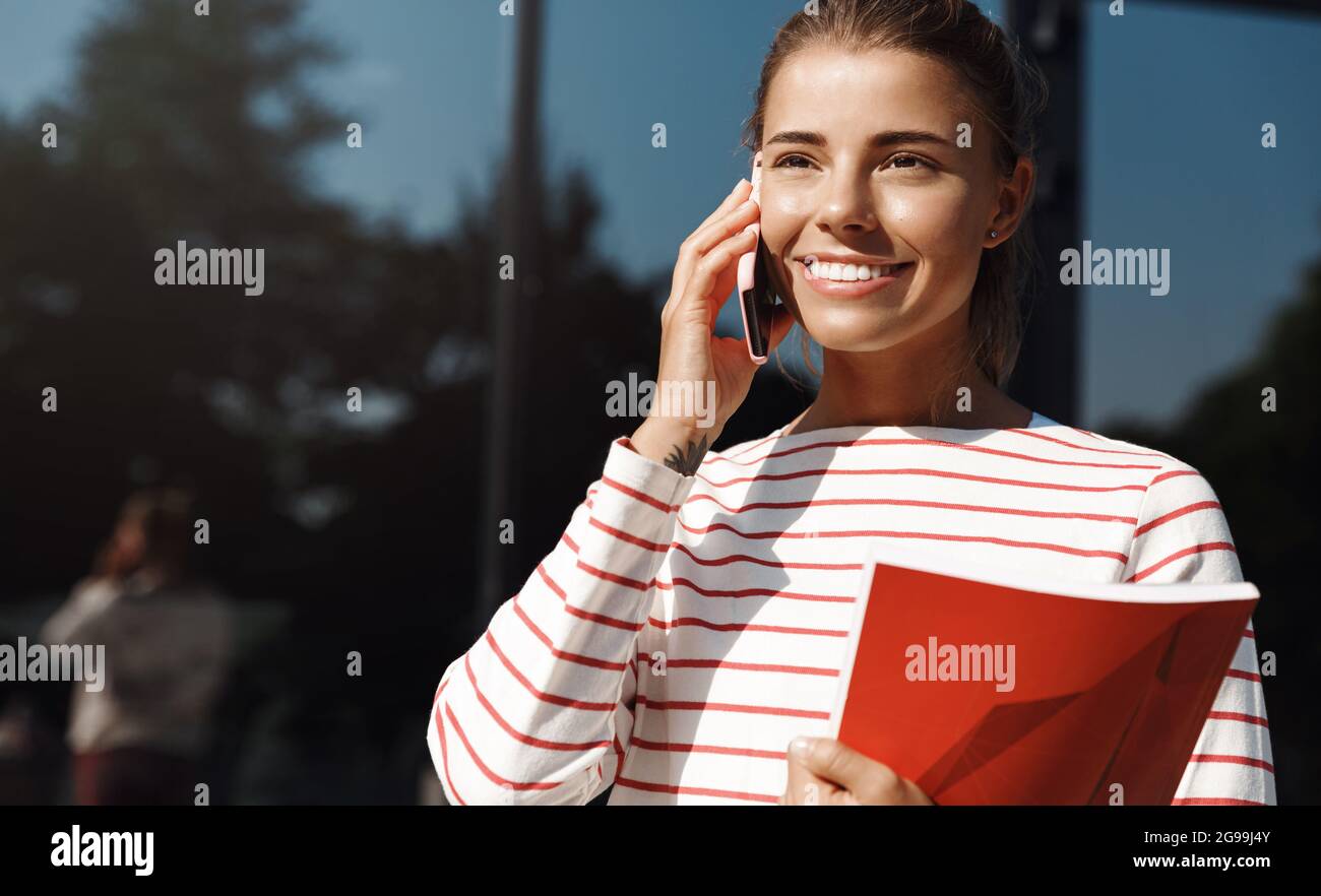 Portrait of smiling girl student, holding college study material, talking on mobile phone outside unviersity, near building. Woman having a call near Stock Photo