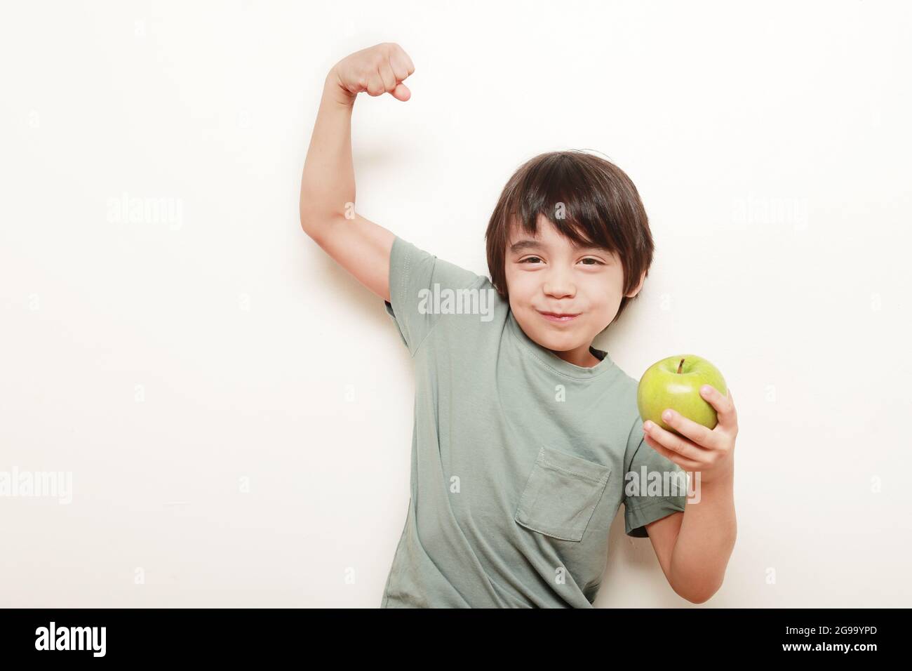 child on a white background eating the green apple he holds in one hand and with the other hand he shows us his bicep indicating strength Stock Photo