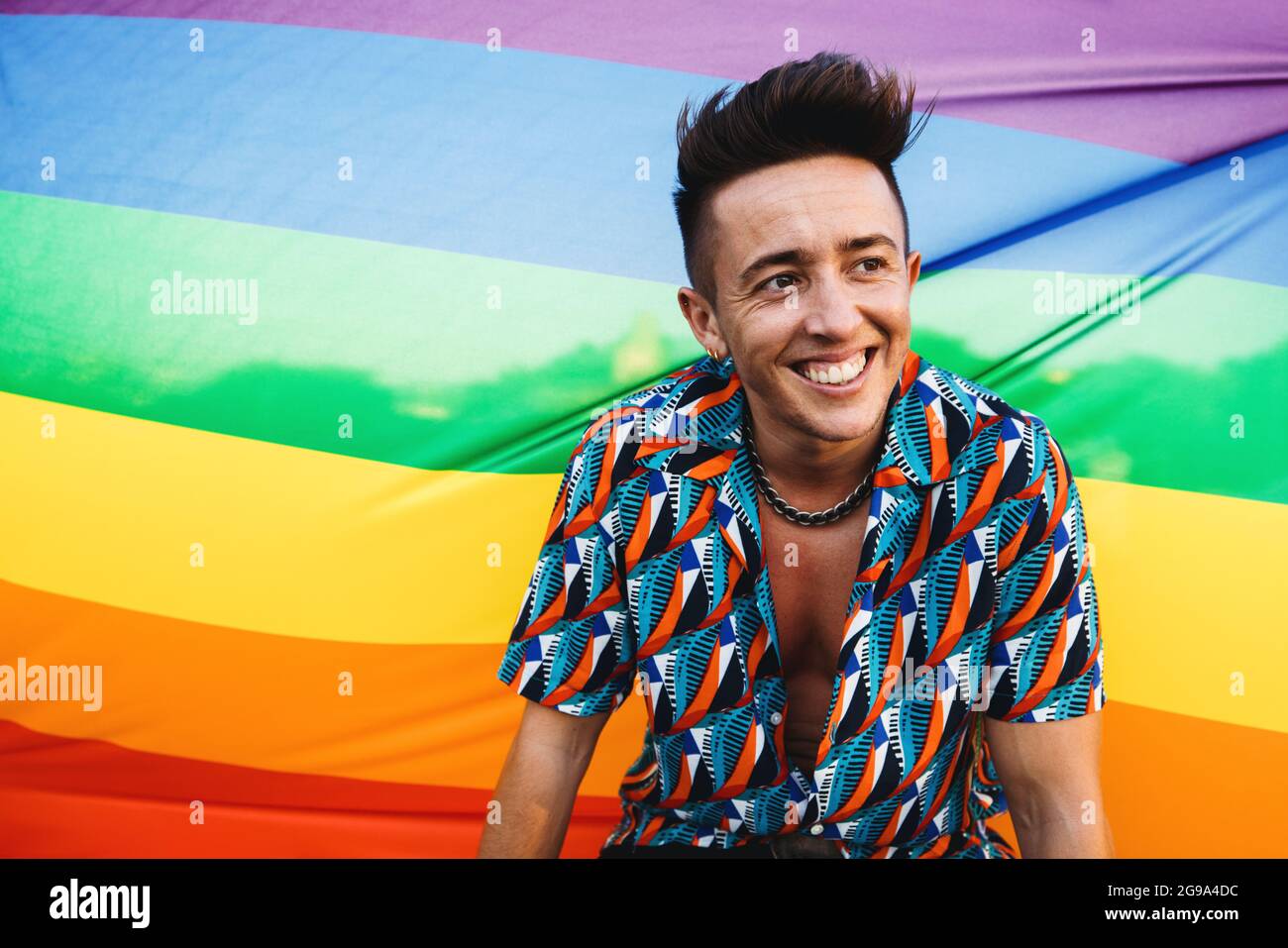 Happy transgender man embracing his queer identity. Young transman smiling cheerfully while standing against a pride flag. Young queer man posing agai Stock Photo