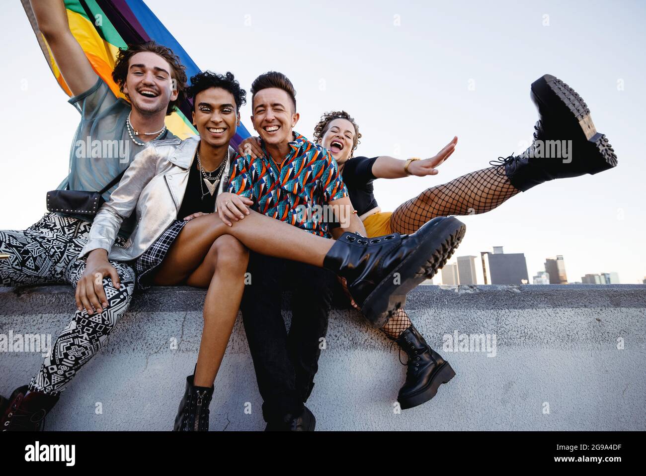 Four LGBTQ people celebrating pride while sitting together. Four friends smiling cheerfully while raising the rainbow pride flag. Group of young queer Stock Photo