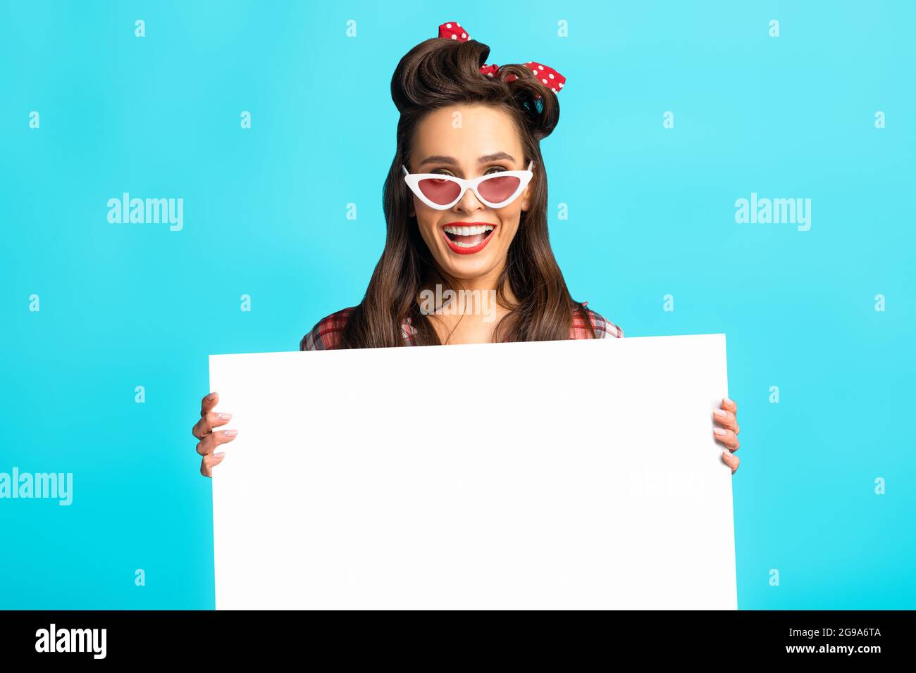 Shocked pinup woman in retro wear presenting something on white blank poster over blue studio background, mockup Stock Photo