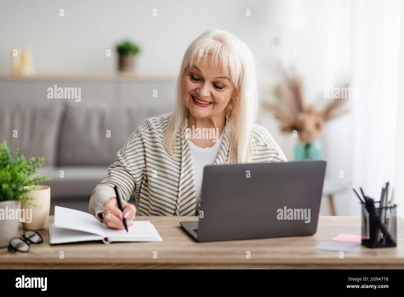 Smiling mature lady writing in notebook using laptop at home Stock Photo