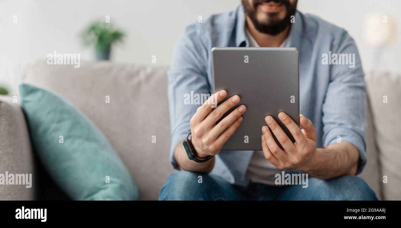 Unrecognizable man sitting on sofa, using nice mobile app on digital tablet, panorama, copy space, crop Stock Photo