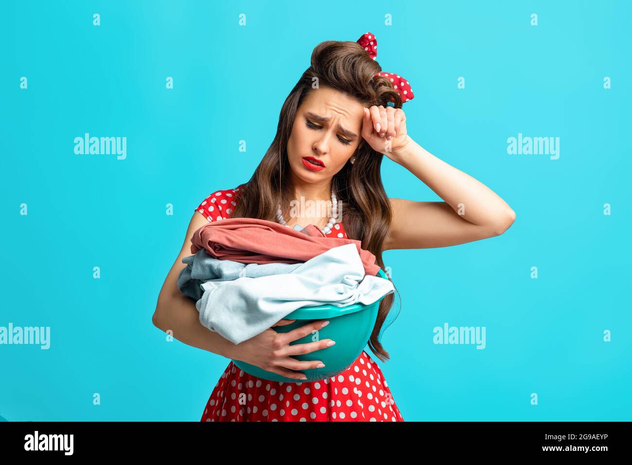 Sad exhausted pinup woman in retro outfit holding clothes for washing or ironing on blue studio background Stock Photo