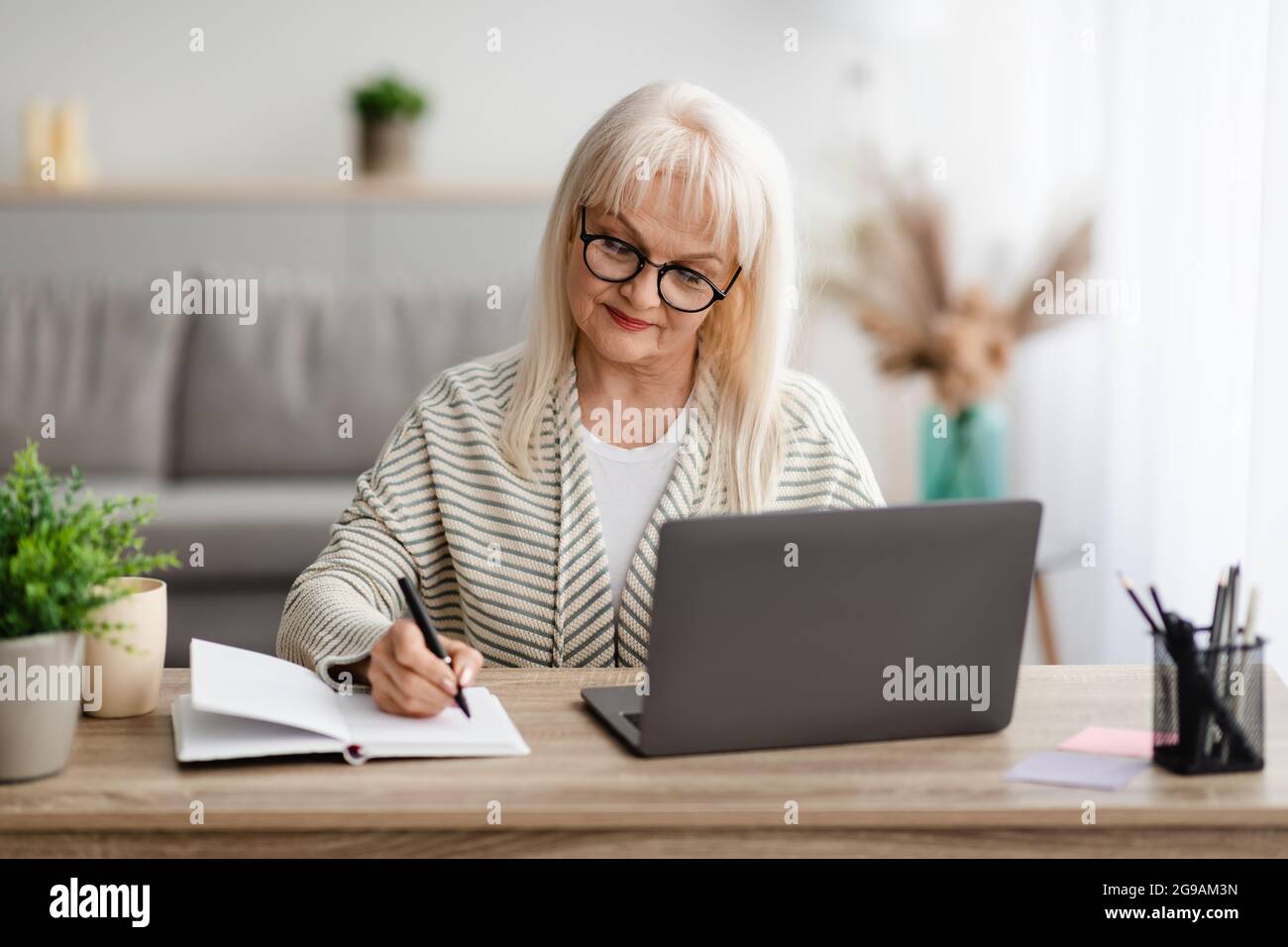 Mature lady writing in notebook and using laptop at home Stock Photo
