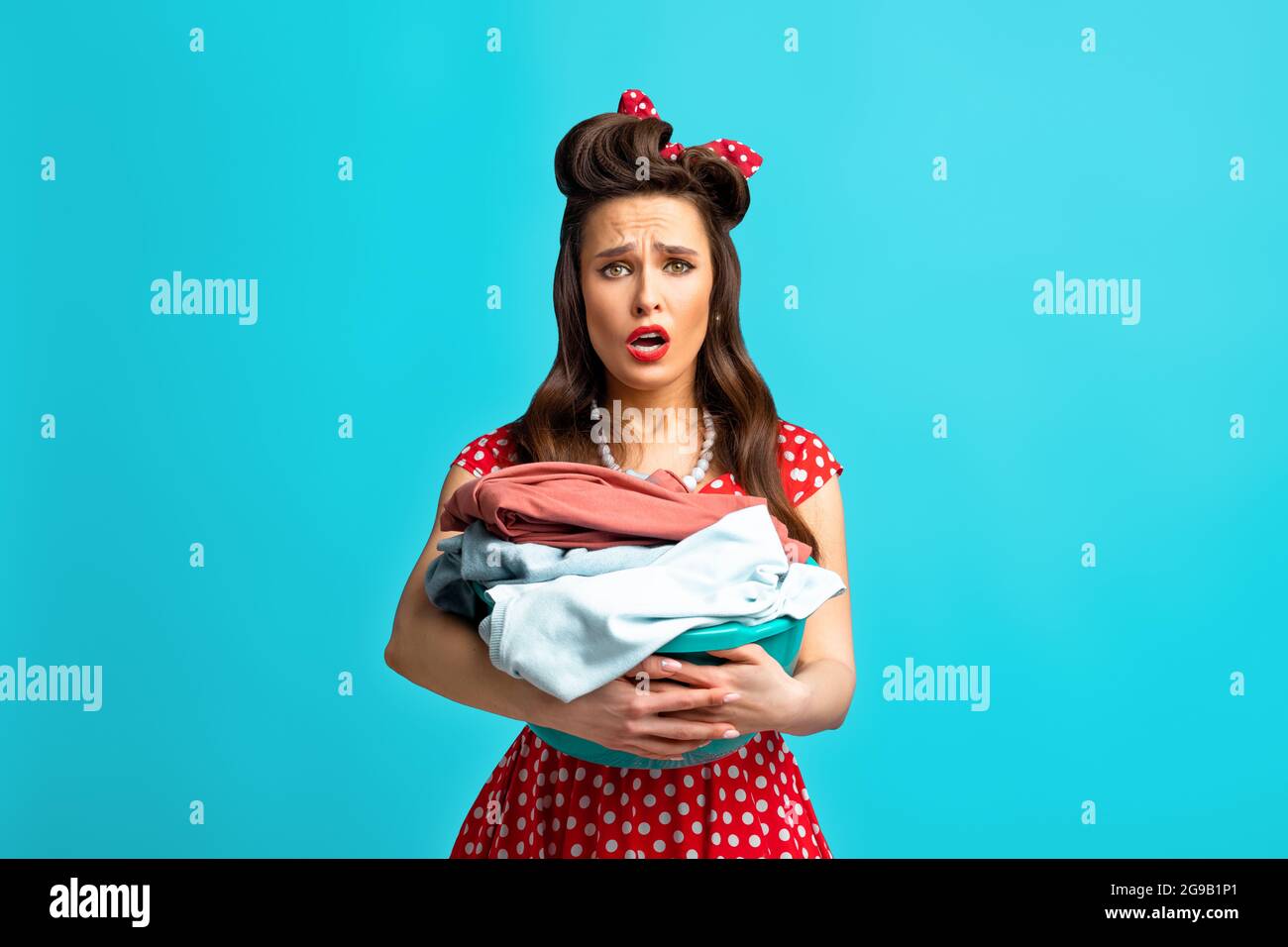 Unhappy tired young pinup woman in retro dress holding basin with laundry for washing or ironing on blue background Stock Photo