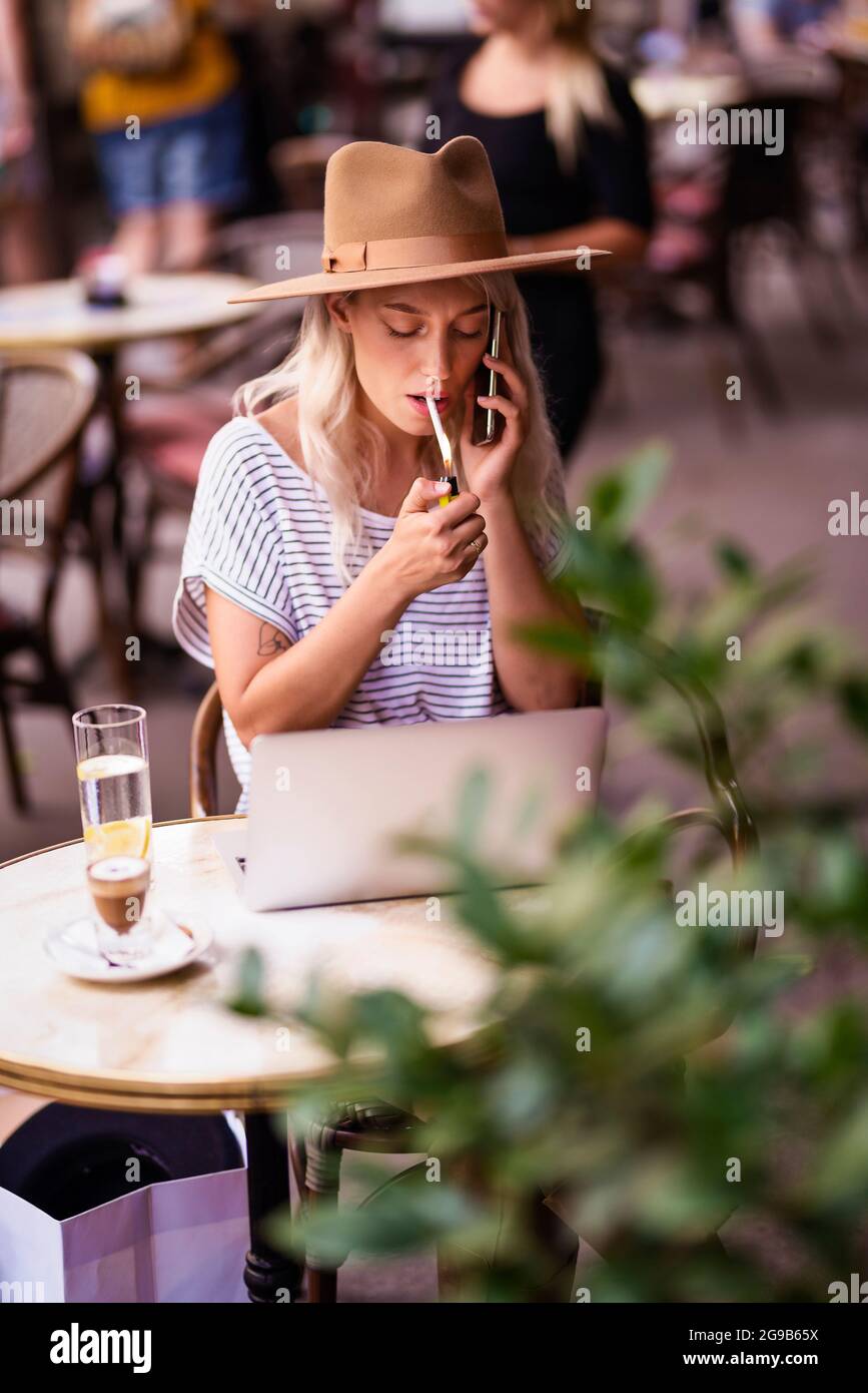 Young woman lighting a cigarette and talking on mobile phone while sitting outdoors at the typical french cafe terrace. Stock Photo