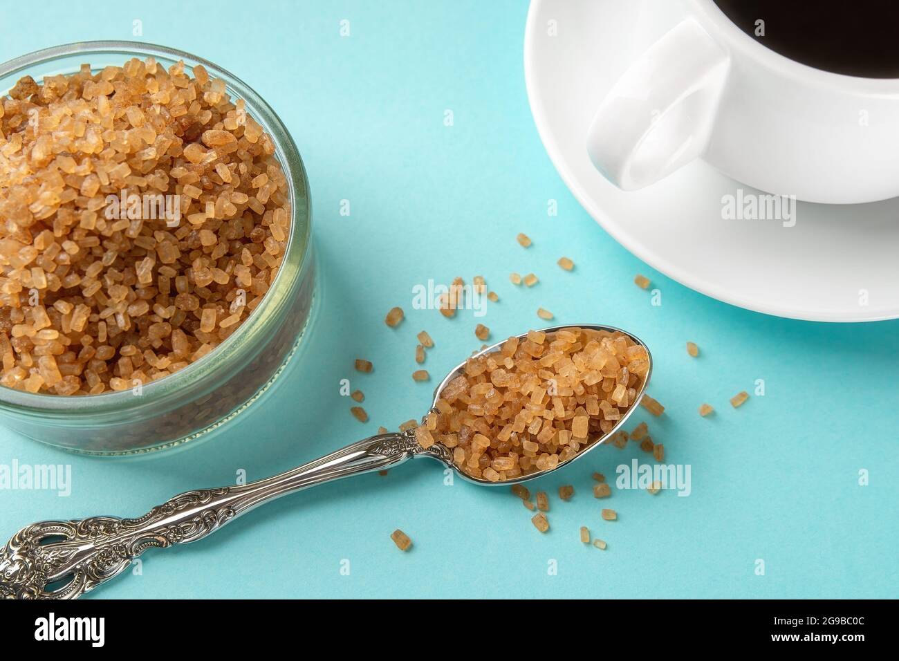 Amber sugar crystals in a spoon near a glass sugar bowl and a cup of hot drink over blue background. Unrefined brown cane sugar for coffee and tea clo Stock Photo