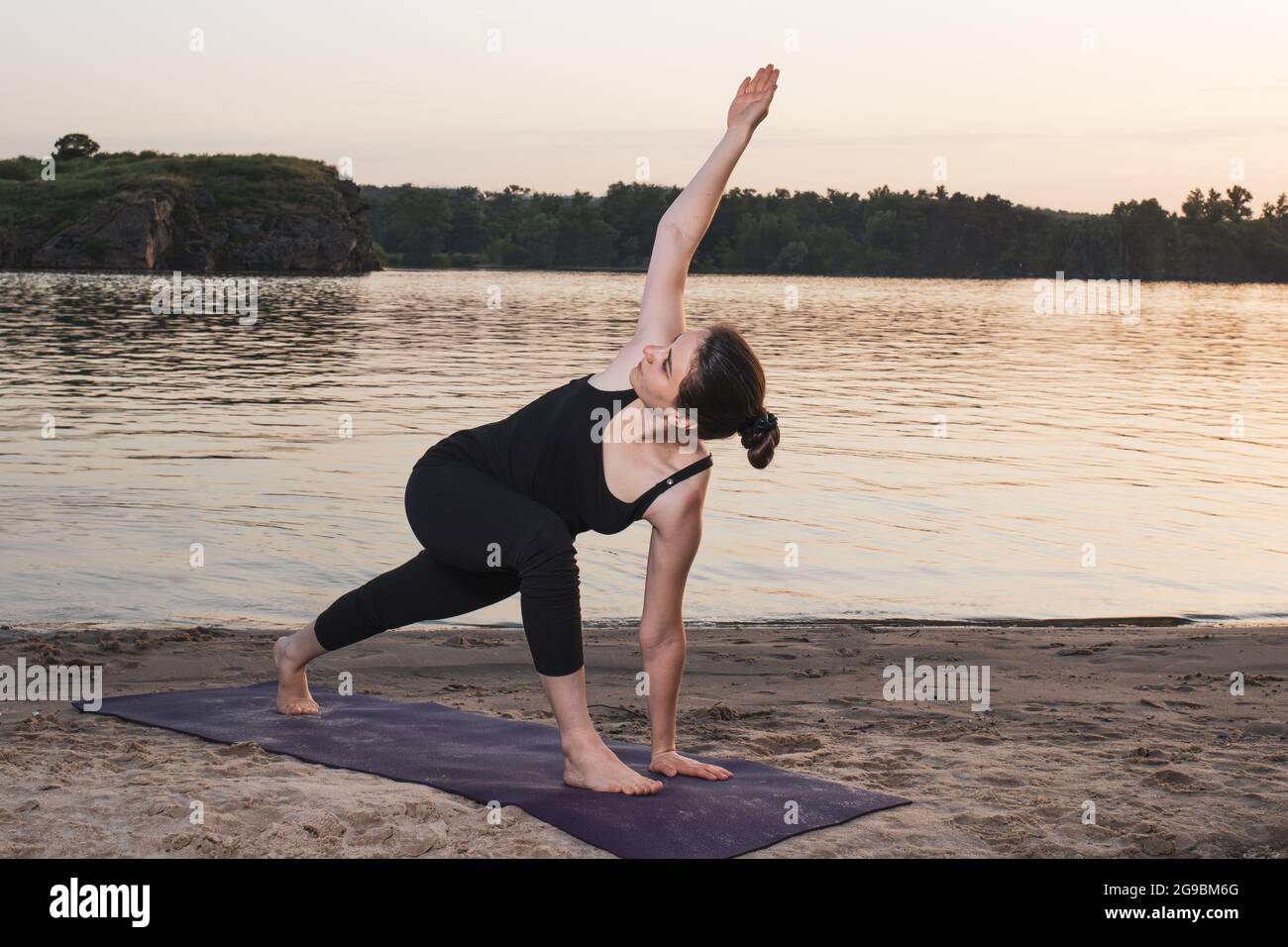 Girl Doing Yoga on a mat on the beach by the river at sunset, twisting in a standing position. Extended Side Angle Pose Utthita Parsva Konasana Stock Photo