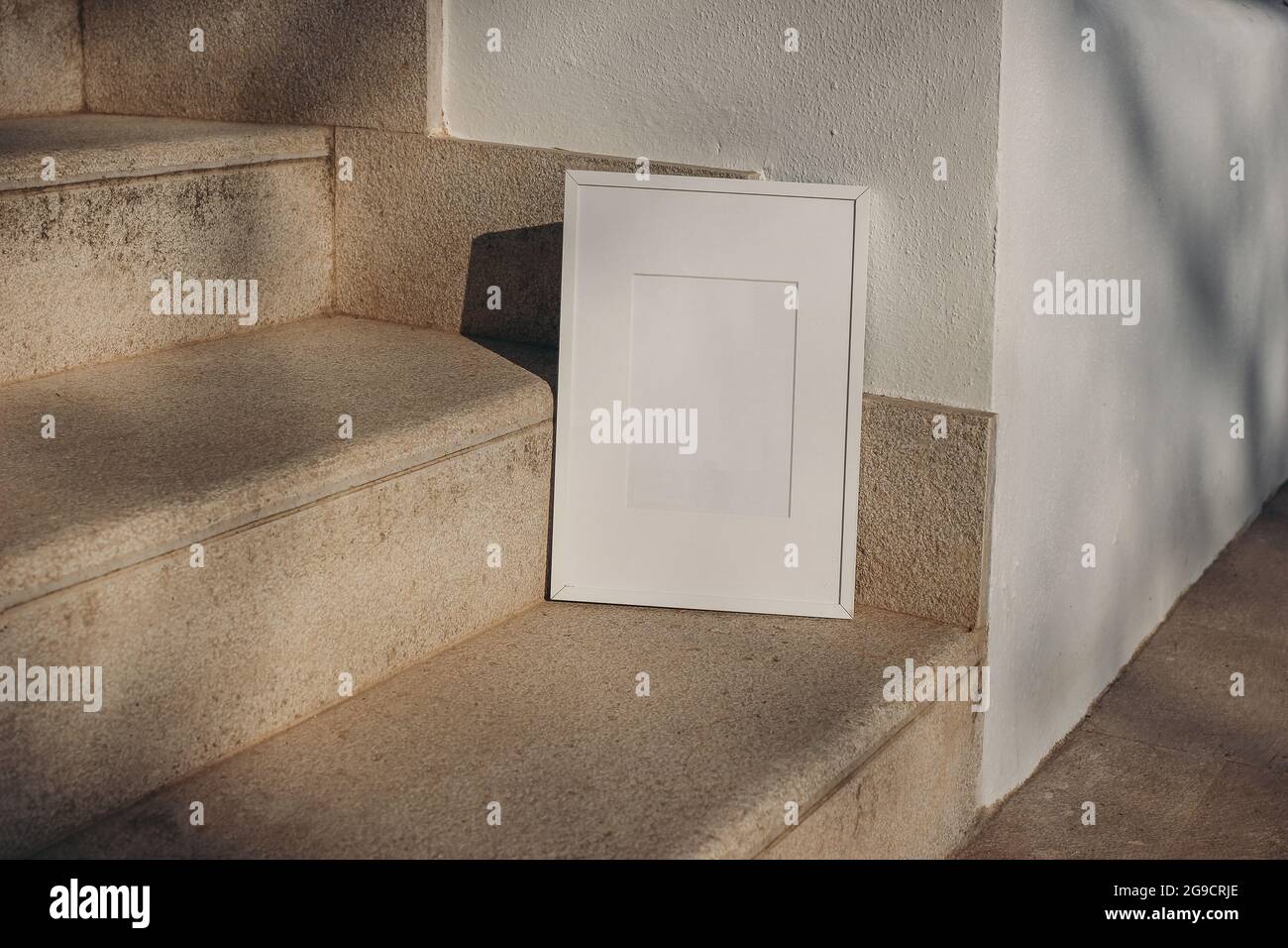 Blank white picture frame leaning against white wall. Outdoor sandstone stairs in sunlight, shadows overlay. Empty poster mockup for art display Stock Photo