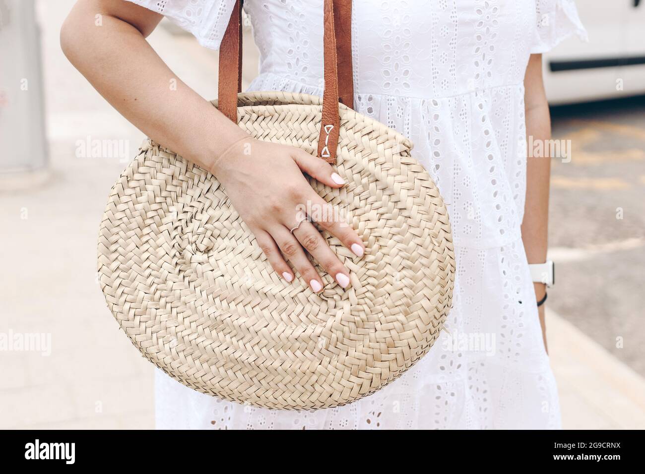 Closeup of young caucasian woman in white madeira lace dress holding round straw bag with leather straps. Hands with pink polish nails and ring Stock Photo