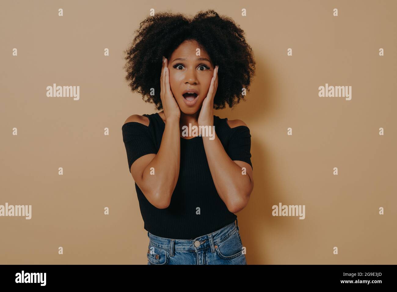 Photo of shocked pretty dark skinned curly haired lady holding hands on cheekbones Stock Photo