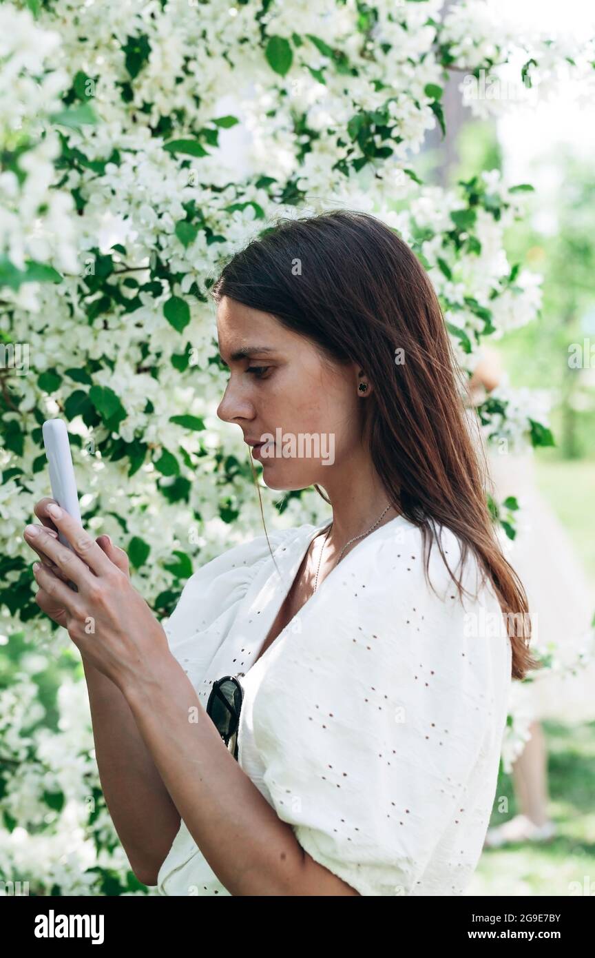 Attractive and beautiful woman on the apple tree background uses mobile technologies. Young woman caucasian ethnicity. Cheerful pretty model with phon Stock Photo