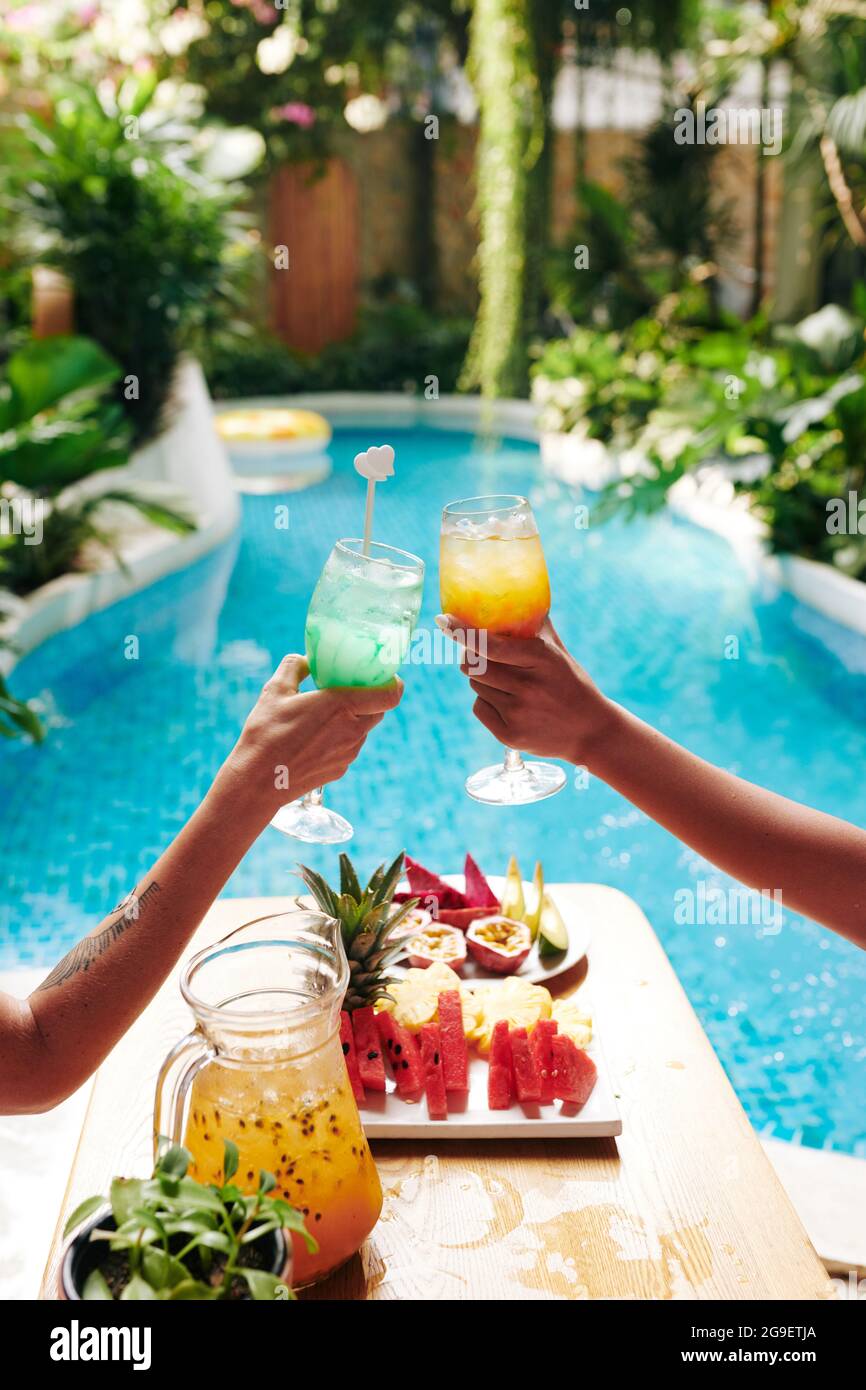 Female friends relaxing in chaiselounges by swimming pool and clinking glasses of delicious