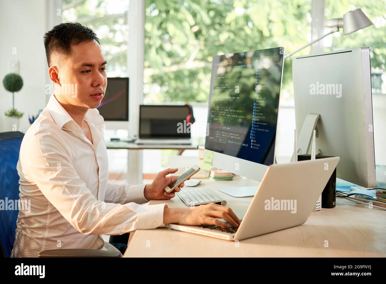 Young software engineer with smartphone in hand working on laptop when checking programming code Stock Photo