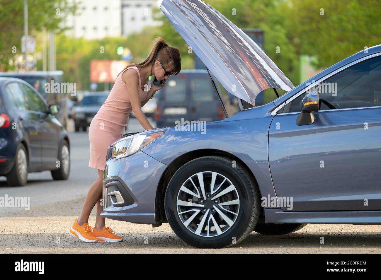 Helpless woman standing near her car with open hood takling on mobile phone calling road service for help. Young female driver having trouble with veh Stock Photo