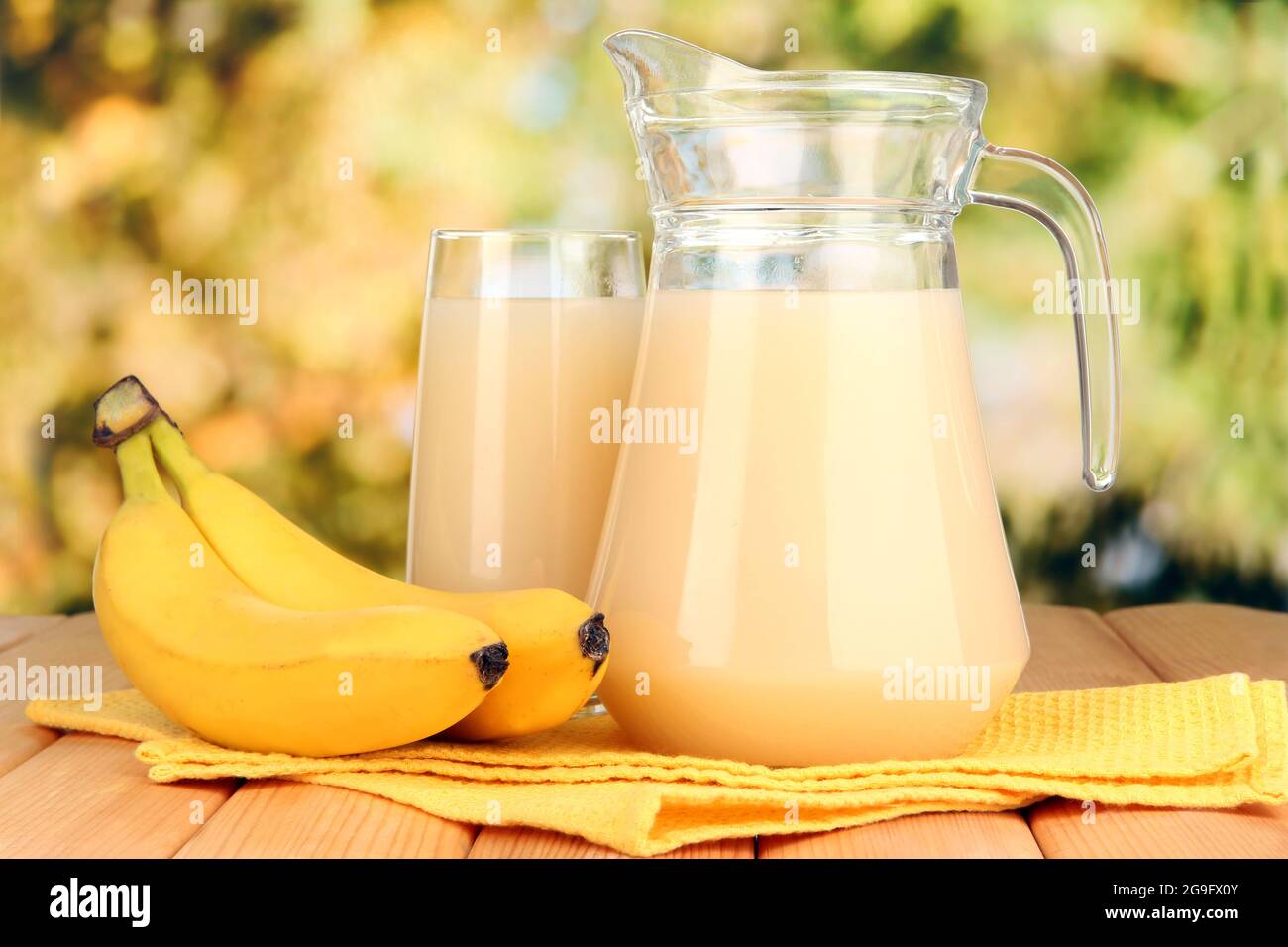 Full glass and jug of banana juice and bananas on wooden table outdoor