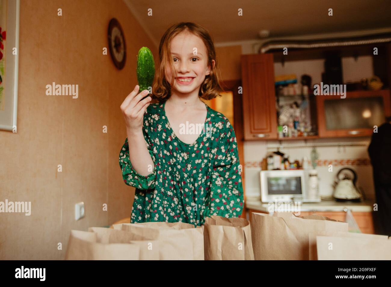 Joyful girl in green dress demonstrating ripe cucumber and looking at camera while standing in kitchen Stock Photo