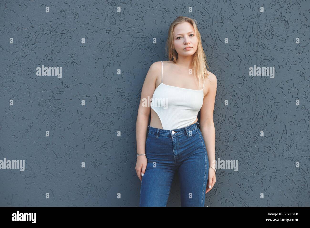 young blonde woman in the white sleeveless t-shirt and blue jeans standing over gray wall background Stock Photo