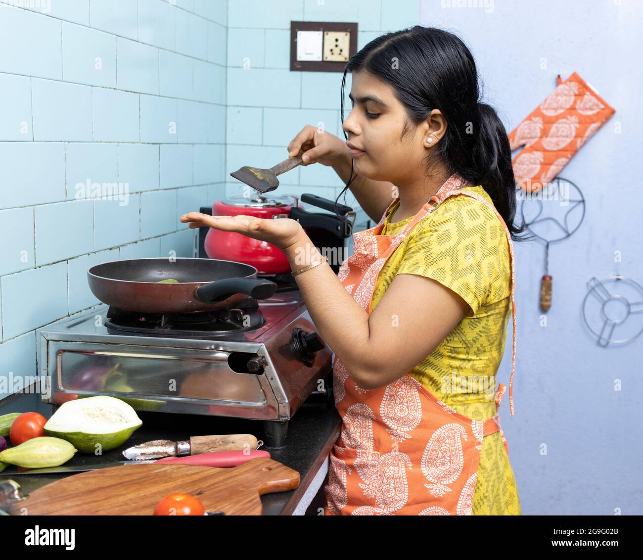 A Pretty Indian Young Woman Wearing Apron Cooking And Tasting Food In 