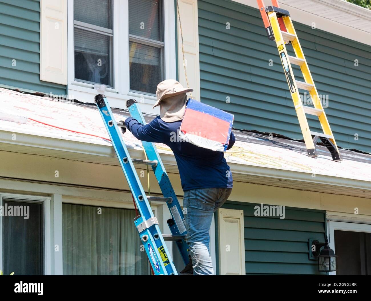 A contractor is carrying a package of roof shingles while climbing a ladder to install on a residential home. Stock Photo