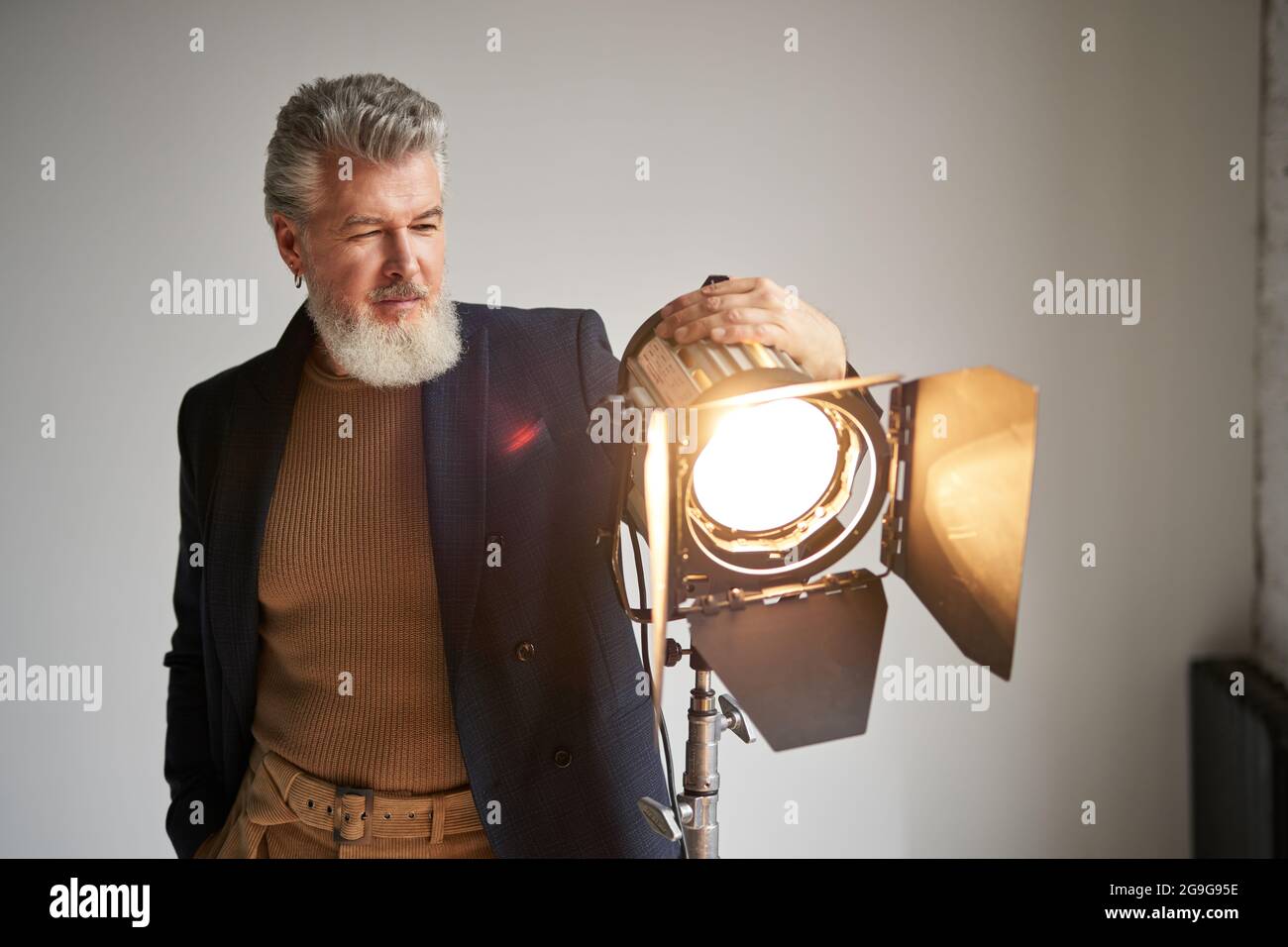Portrait of handsome bearded middle aged man dressed elegantly standing next to studio spotlight while posing for camera over white background Stock Photo
