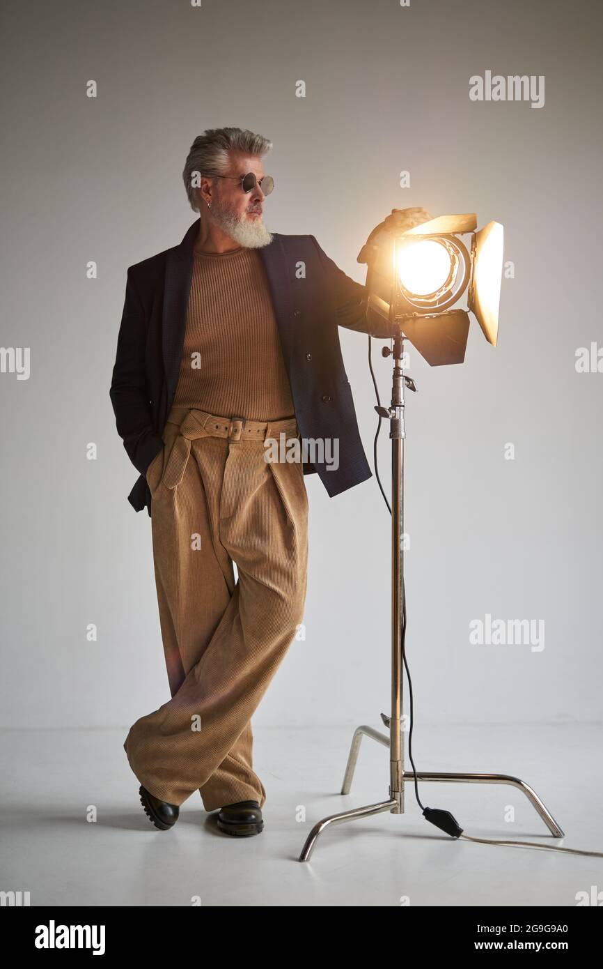 Full length shot of trendy bearded middle aged man in glasses looking aside, standing next to studio spotlight while posing for camera over white Stock Photo
