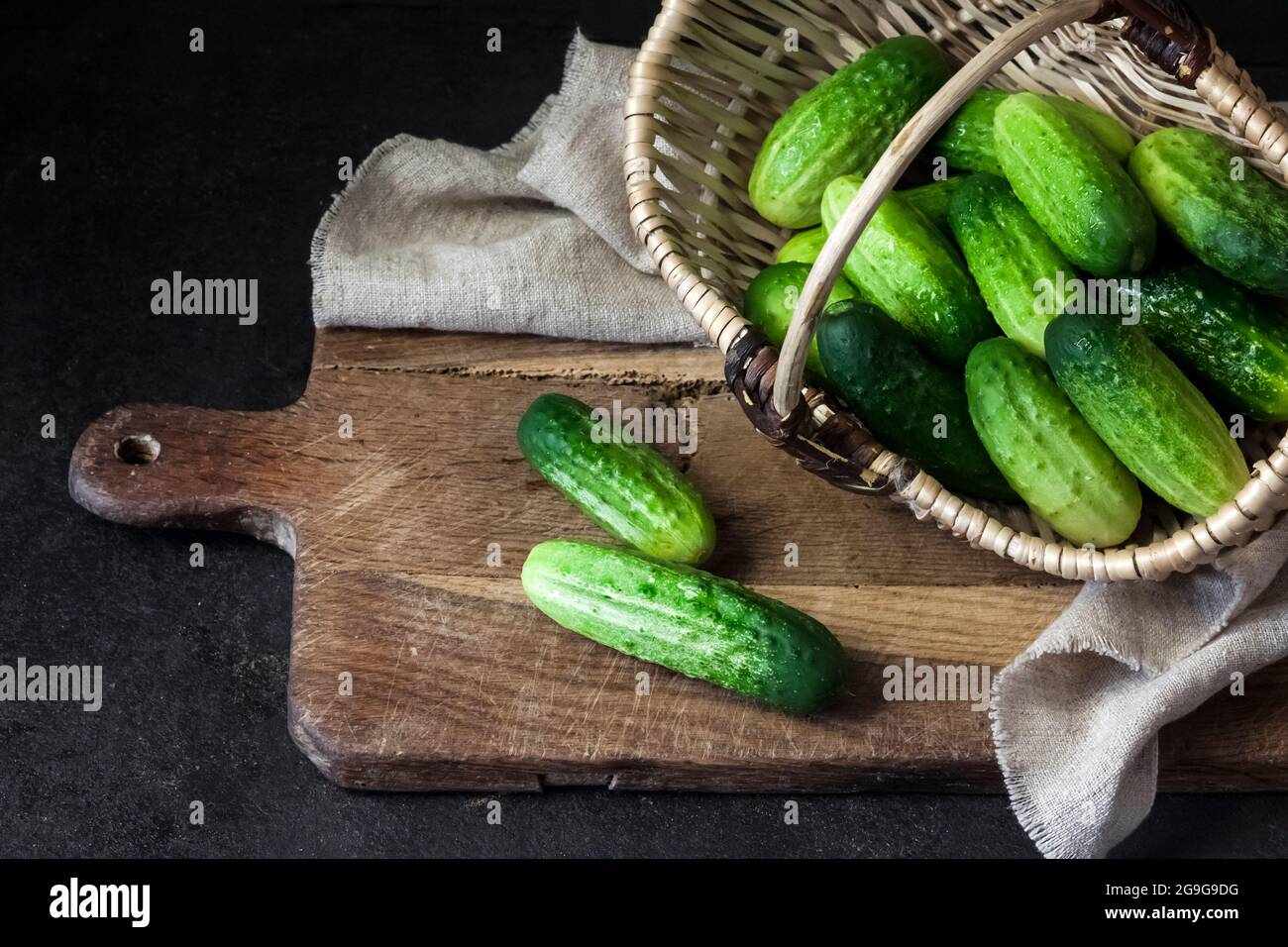 Fresh cucumbers in wicker basket on wooden board on black background. Stock Photo