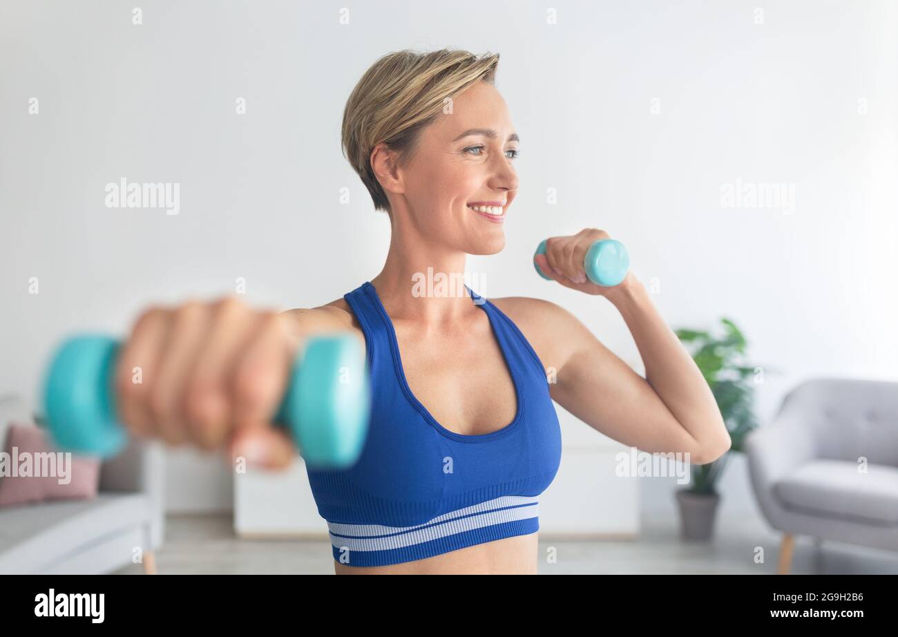 Domestic Fitness Concept. Portrait Of Smiling Adult Woman In Sports Bra  Exercising With Two Dumbbells Posing At Home, Enjoying Workout, Looking  Away Stock Photo - Alamy