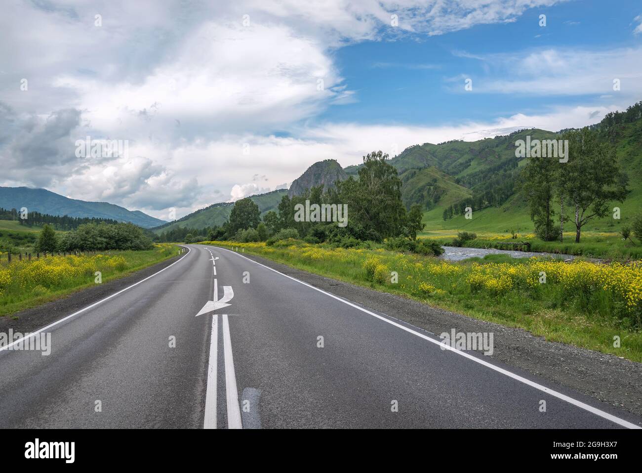 Amazing view with an asphalt road in the mountains, a river, trees and bright yellow flowers on the roadside against the backdrop of blue sky and clou Stock Photo