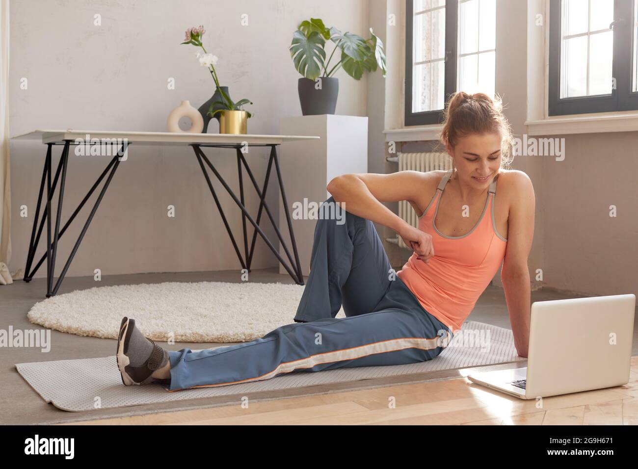 Smiling athletic female in sportswear sitting on mat with laptop during online workout at home and looking at screen Stock Photo