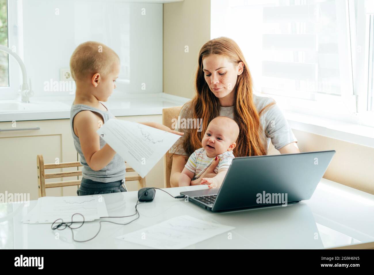 Young attractive mother with two children sitting at the table in the kitchen at home and works on a laptop while taking care of the children. Stock Photo
