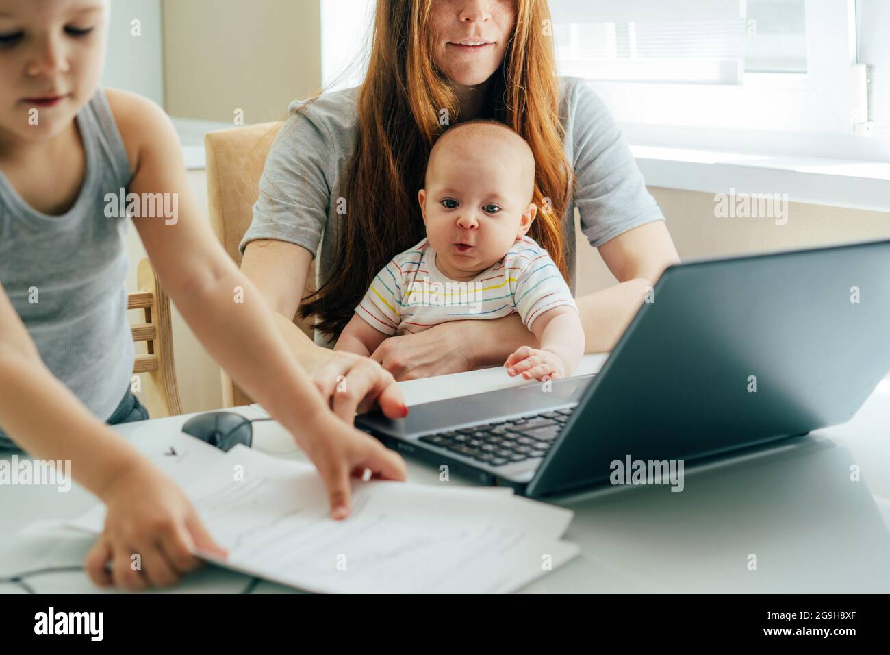Young focused business mom with two children working remotely on a laptop at home office. Mom holds the baby on her lap. Stock Photo