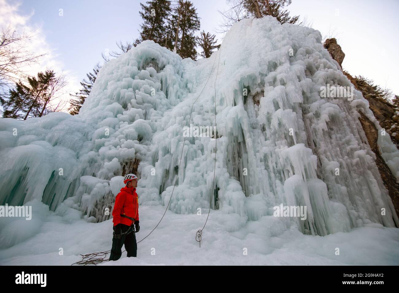 Man standing and controlling a safety top rope while female with ice climbing equipment, climbing on a frozen waterfall Stock Photo