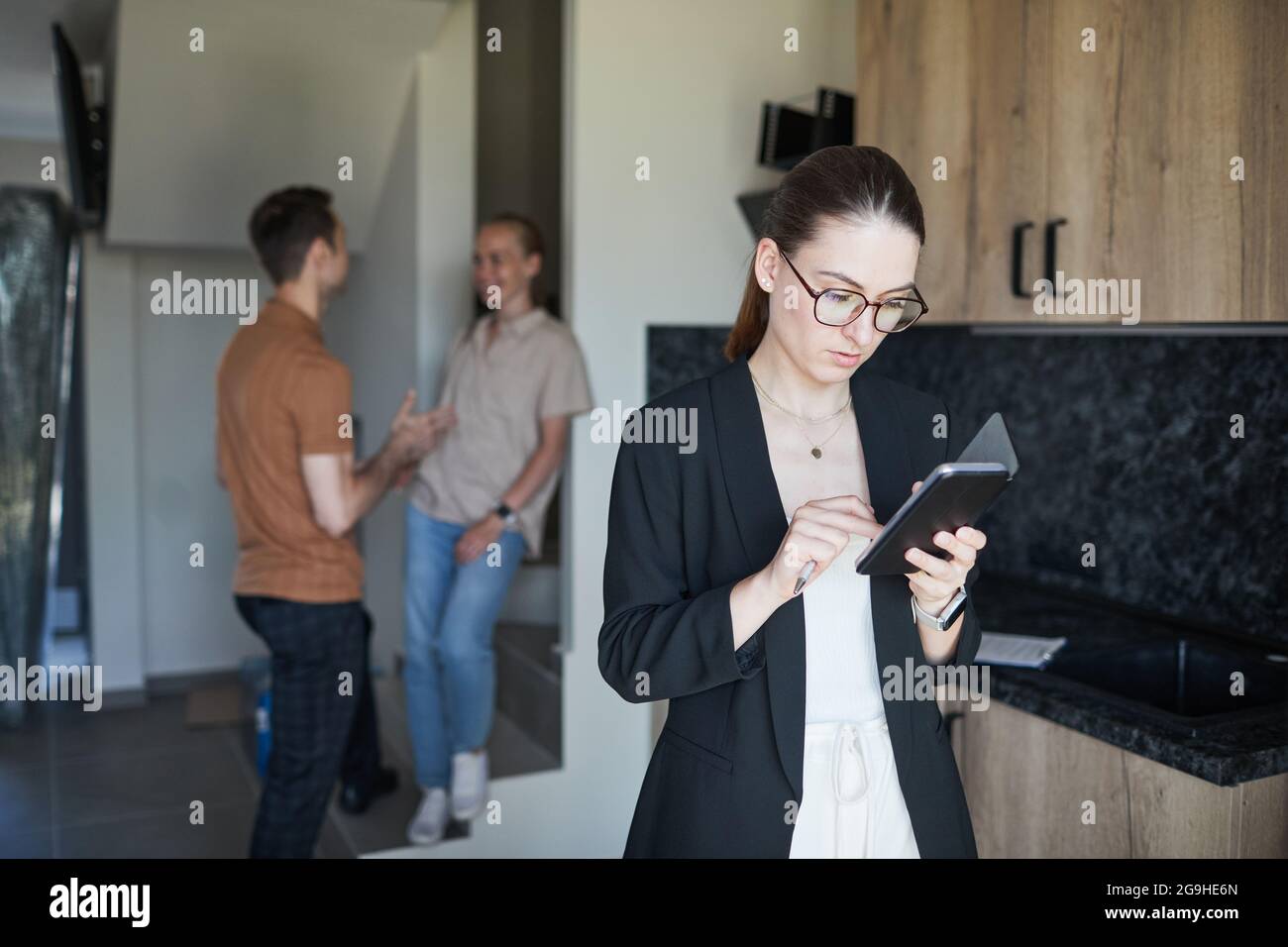 Waist up portrait of female real estate agent using smartphone with young couple in background, copy space Stock Photo