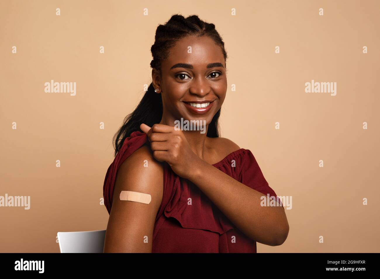 Closeup of pretty black lady showing medical band on her shoulder after vaccination and smiling at camera, beige studio background, copy space. Immuni Stock Photo