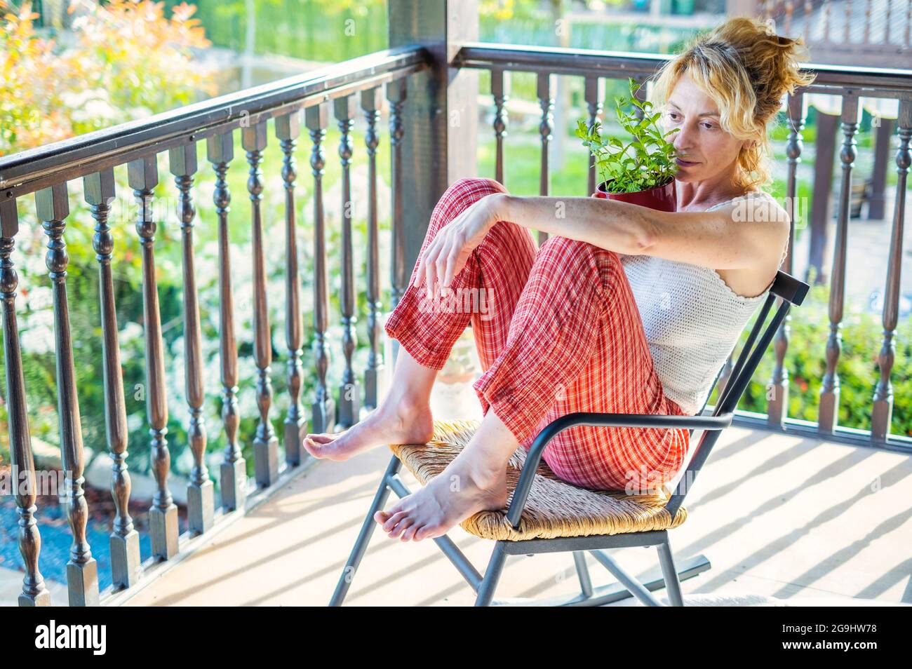 portrait of a young mature caucasian blonde woman relaxing at home on the porch sitting in a rocking chair with a potted plant. Lifestyle concept. Stock Photo