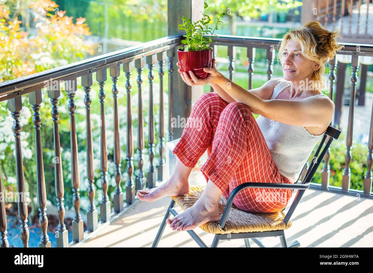 portrait of a young mature caucasian blonde woman relaxing at home on the porch sitting in a rocking chair with a potted plant. Lifestyle concept. Stock Photo