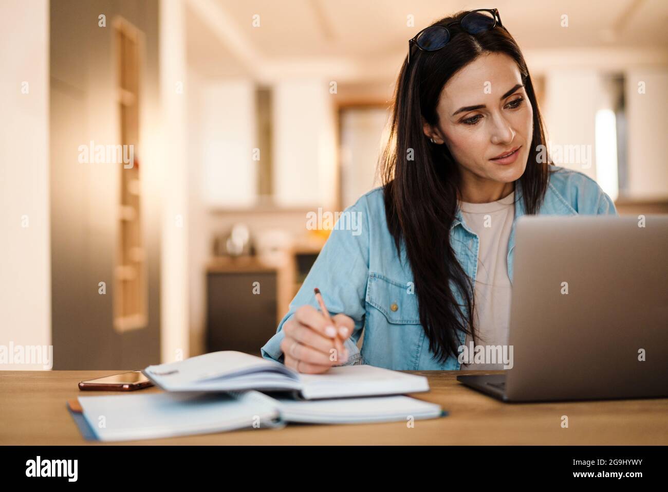 Focused charming woman writing down notes while working with laptop at ...