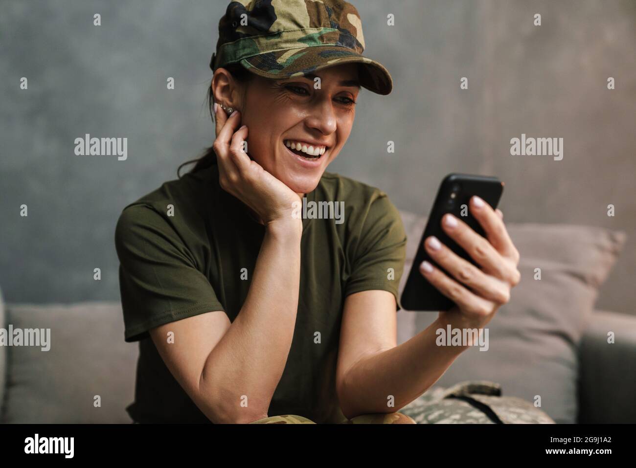Smiling beautiful soldier woman using mobile phone while sitting on sofa indoors Stock Photo