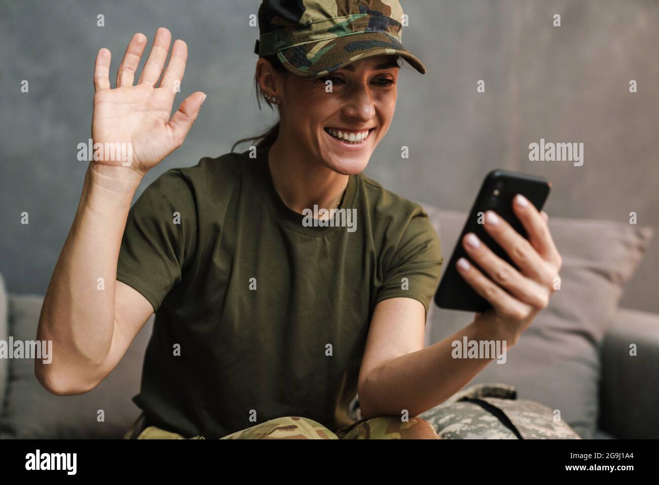 Smiling beautiful soldier woman waving hand while using mobile phone indoors Stock Photo