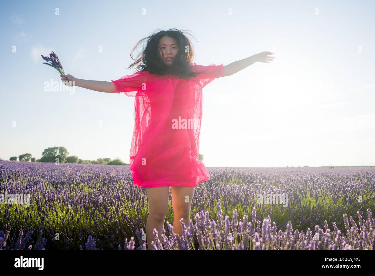 young Asian woman outdoors at lavender flowers field - happy and beautiful Chinese girl in sweet Summer magenta dress enjoying holidays relaxed on pur Stock Photo