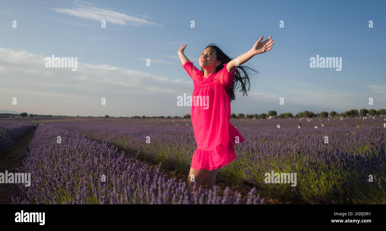 young Asian woman outdoors at lavender flowers field - happy and beautiful Japanese girl in sweet Summer magenta dress enjoying holidays relaxed on pu Stock Photo