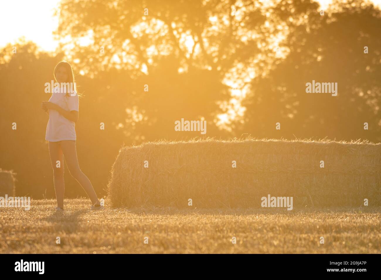 Teenage girl with long blonde hair wearing shorts and white t-shirt posing standing in a field with straw bales. Warm atmosphere due to the falling su Stock Photo