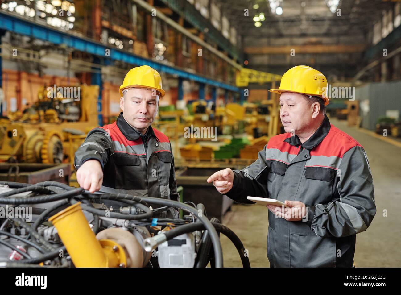 Two workers in helmets using tablet pc and discussing the work of machine equipment in the plant Stock Photo