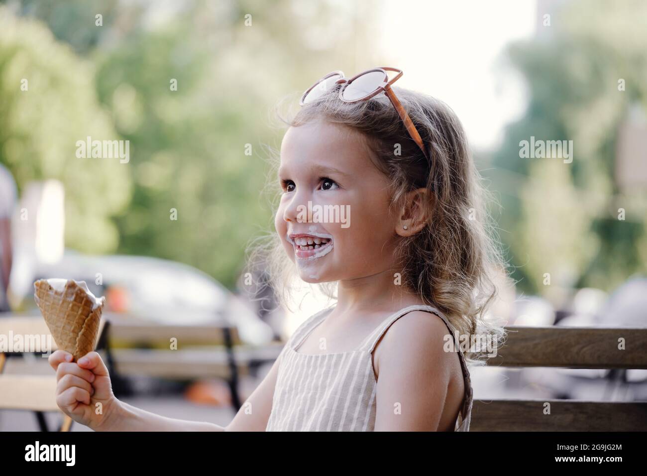 Toddler child eating cone ice cream outside near cafe. Cute blonde Caucasian girl licking frozen summer food on the street. Dirty mouth, happy kid. Stock Photo